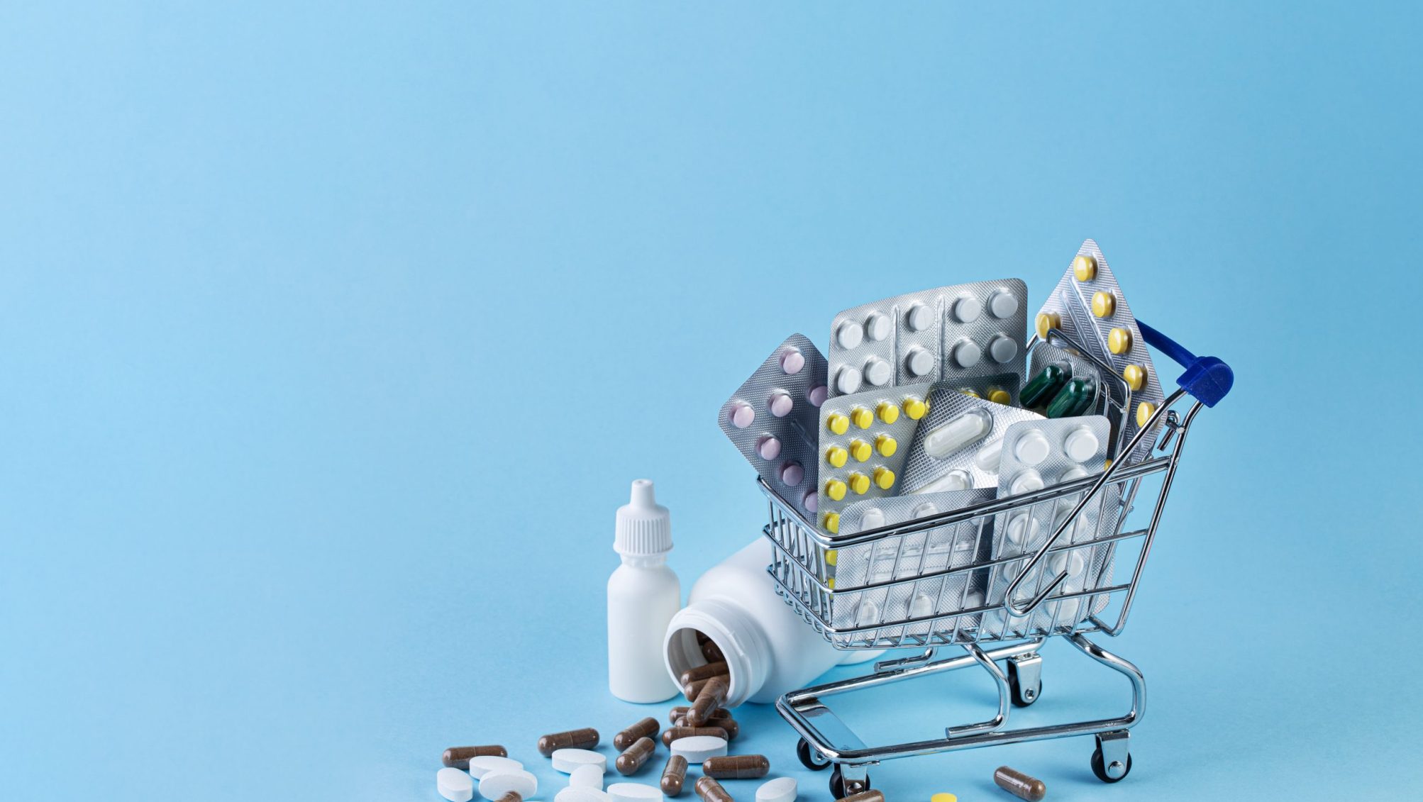 Various capsules, tablets and medicine in shop trolley and pills spilling out of white bottle on blue background. Buy and shopping medicine concept.